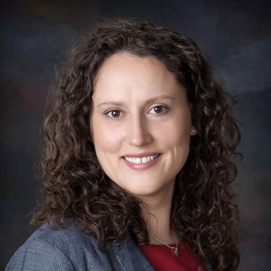 A person with curly brown hair and a navy blazer smiles at the camera. The background is a dark gradient with subtle shades of blue and gray.